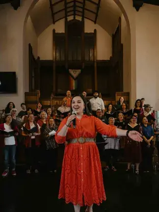Natalia Romero standing in front of chorus with microphone in hand smiling and left arm outstretched